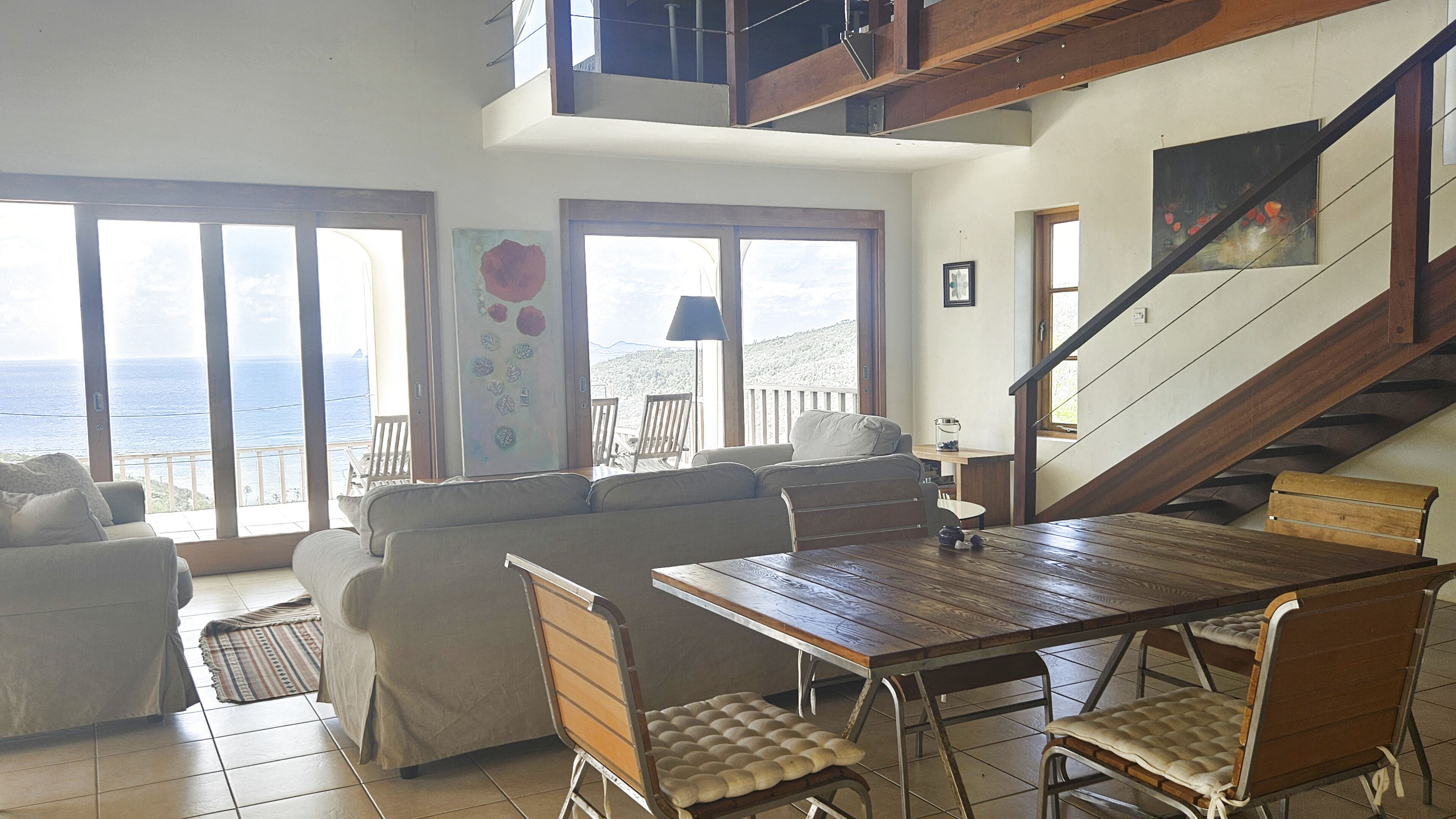 A spacious living area with a wooden dining table, cushioned chairs, a sofa, and a view of the ocean through large windows.