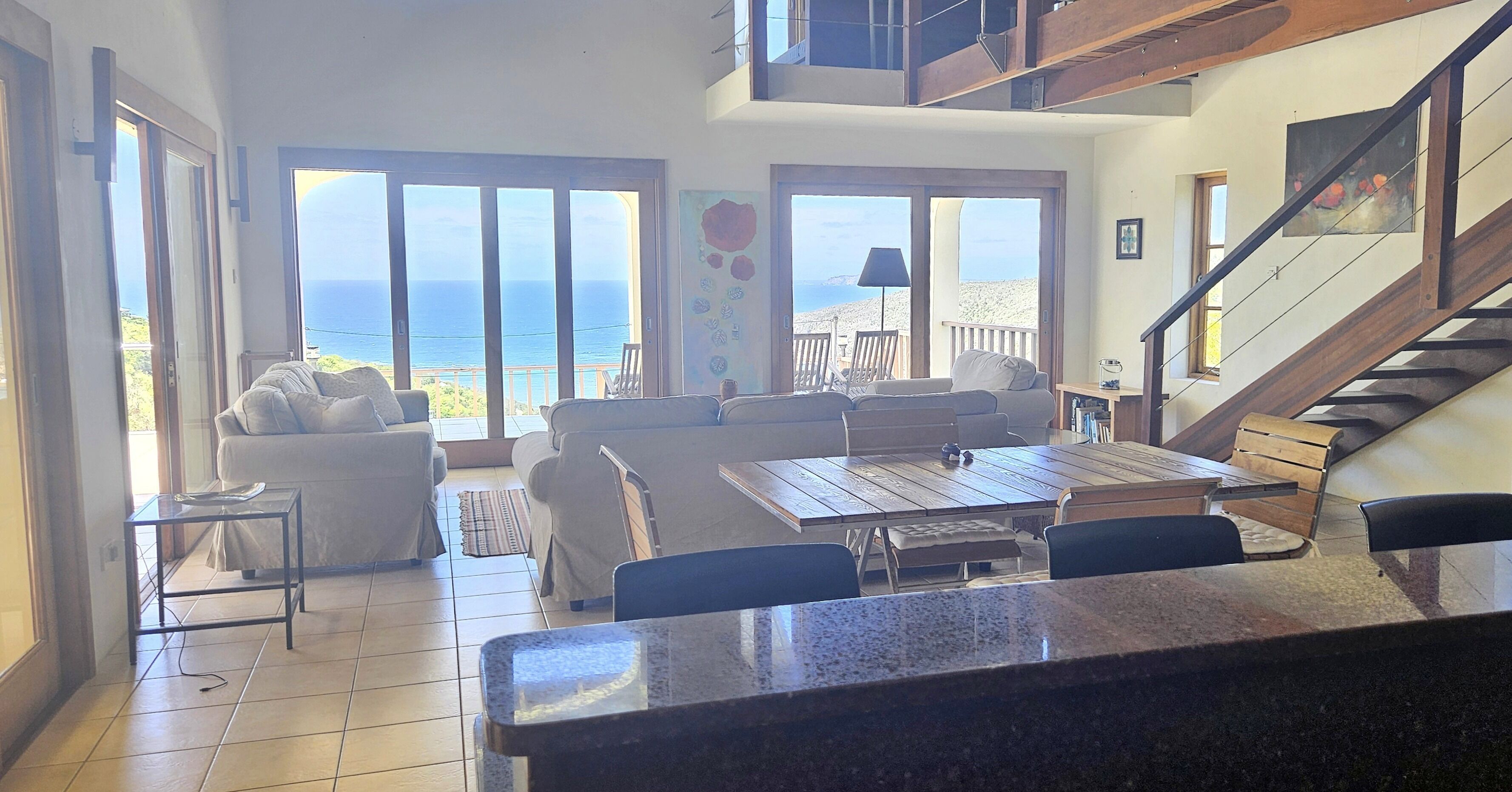 A spacious living area with a view of the ocean, a dining table, and a staircase leading to an upper level.