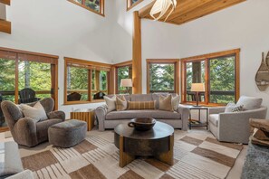 A spacious living room with a large sofa, two armchairs, and a coffee table. There are multiple windows providing views of the outdoors. - Mountain view townhome w/ ski access & hot tub (Whistler)