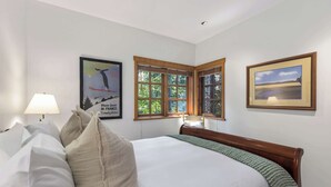 Quarto de hotel com uma cama grande, uma mesa de cabeceira, uma luminária, uma janela com persianas e duas molduras com quadros na parede.