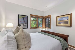 A hotel room with a large bed, a nightstand, a lamp, a window with blinds, and two framed pictures on the wall. - Mountain view townhome w/ pool & hot tub (Whistler)