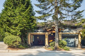 A modern two-story house with a large garage, surrounded by lush greenery and tall trees. - Mountain view townhome w/ ski access & hot tub (Whistler)