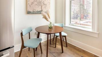 A dining area with a round wooden table, two light blue chairs, a vase with pampas grass, and a painting on the wall.