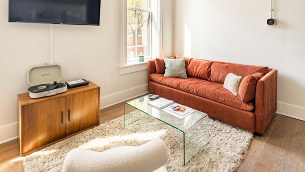 Ein Wohnzimmer mit einem an der Wand befestigten Flachbildfernseher, einer Holzeinrichtung mit Plattenspieler, einem Glastisch, einem roten Sofa und einem weißen, flauschigen Teppich.