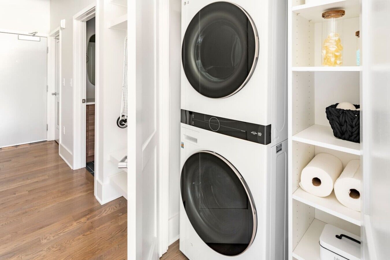 A laundry room with two front-loading washing machines, a shelf with rolls of toilet paper, and a basket on the shelf.