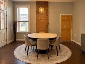 A dining area with a round table, six chairs, a hanging light fixture, a brick accent wall, and a wooden door. - Lux Historic 2BR near Stadiums, Downtown (Cincinnati)