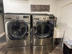 Un área de lavandería con dos lavadoras y secadoras de acero inoxidable de carga frontal, un letrero que dice 'Laundry' en la pared y un lavamanos pequeño con un cesto de basura.
