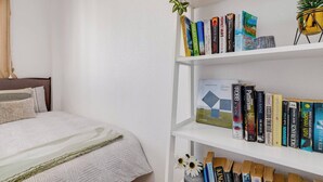 Een slaapkamer met een net opgemaakt bed, een witte boekenkast vol boeken en een kleine plant in een potje op de plank.