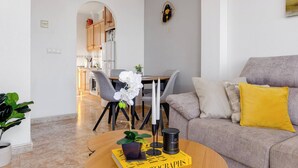 Een moderne woonkamer met een bank, een bijzettafeltje met een boek, en een eethoek op de achtergrond.