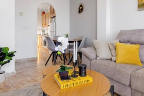 A modern living room with a sofa, a coffee table with a book, and a dining area in the background. - CB Living - Homely apartment Ciñuelica, Punta Prim (Alicante / Alacant)