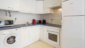 Una cucina compatta con mobili bianchi, lavatrice, microonde, tostapane e frigorifero.
