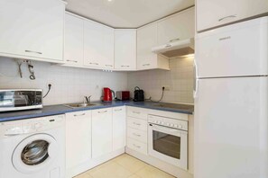 A compact kitchen with white cabinets, a washing machine, a microwave, a toaster, and a refrigerator. - CB Living - Apartment with roof terrace Aguamarina (Orihuela)