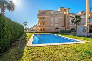 A swimming pool with a surrounding grassy area, a building with balconies, and a clear sky. - CB Living - Apartment with roof terrace Aguamarina (Orihuela)