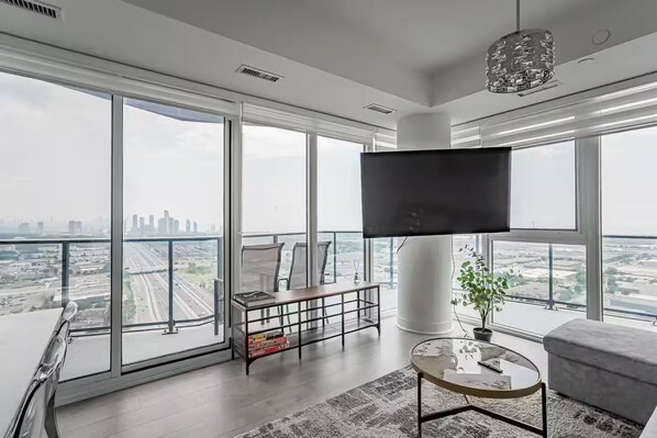 A modern living room with a large TV, comfortable seating, and expansive windows offering city views.