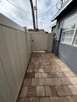 Ein gepflasterter Patio-Bereich mit weißem Zaun, einem Ziegelweg und einem Gebäude mit Fenster und Mauerwerk.