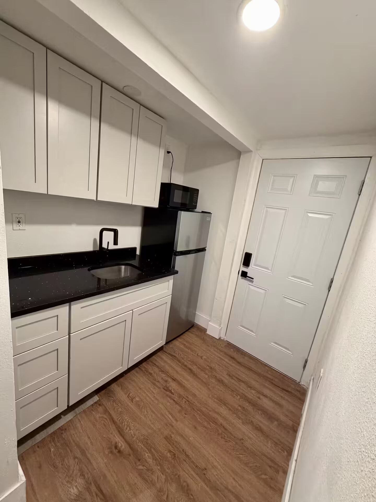 A compact kitchen with white cabinets, a black countertop, a stainless steel sink, a microwave, and a refrigerator.