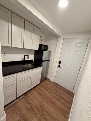 Eine kleine Küche mit weißen Schränken, einer schwarzen Arbeitsplatte, einer Edelstahlspüle, einer Mikrowelle und einem Kühlschrank.