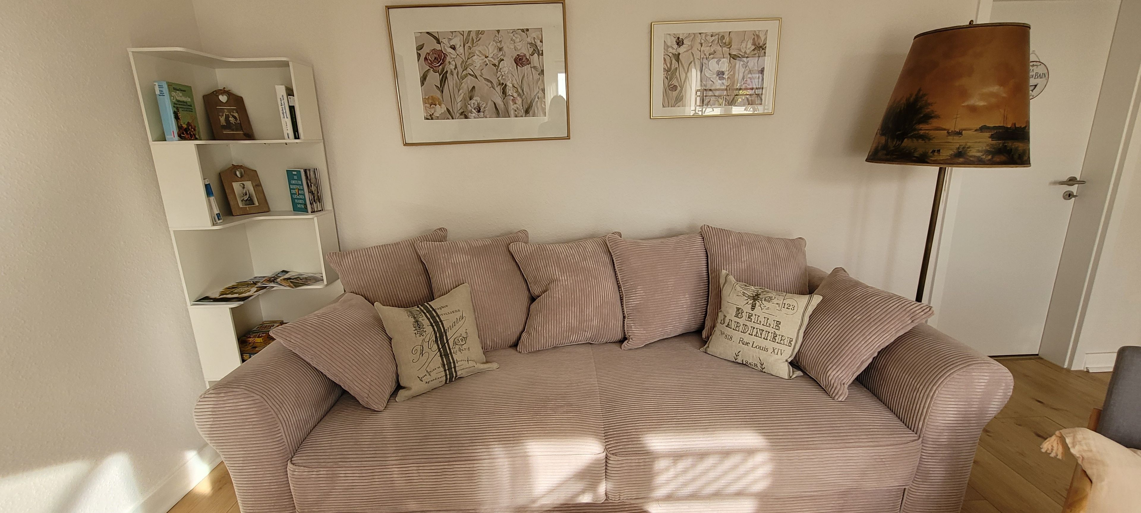 A living room with a beige sofa, striped cushions, a bookshelf, and two framed floral paintings.