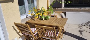 Ein Holztisch mit Stühlen steht auf einer Terrasse mit einer Blumenkomposition.