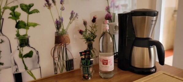 A coffee maker, a bottle of Naturell, and a glass jar with green plants on a wooden surface.
