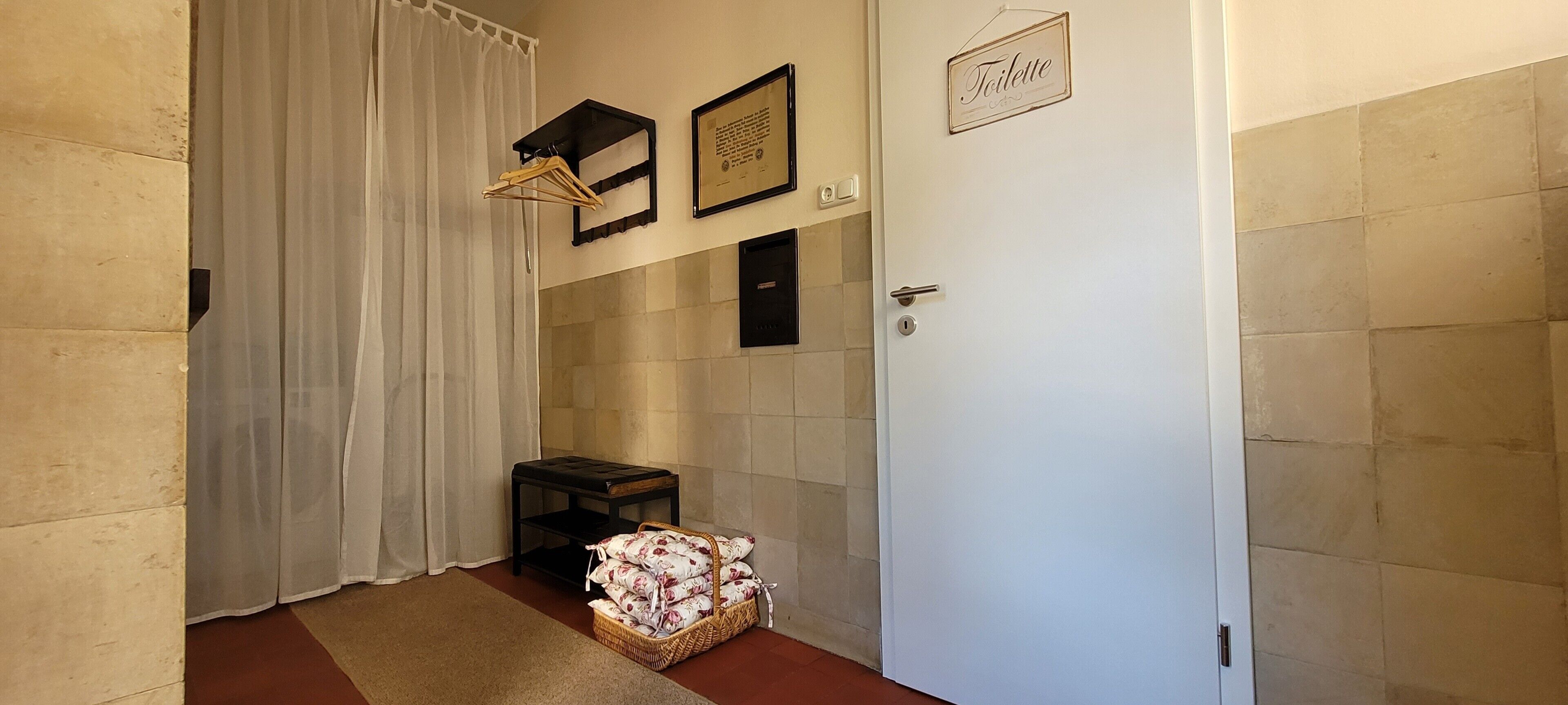 A hallway with a white door, a framed certificate on the wall, a small table with a basket, and a white curtain.