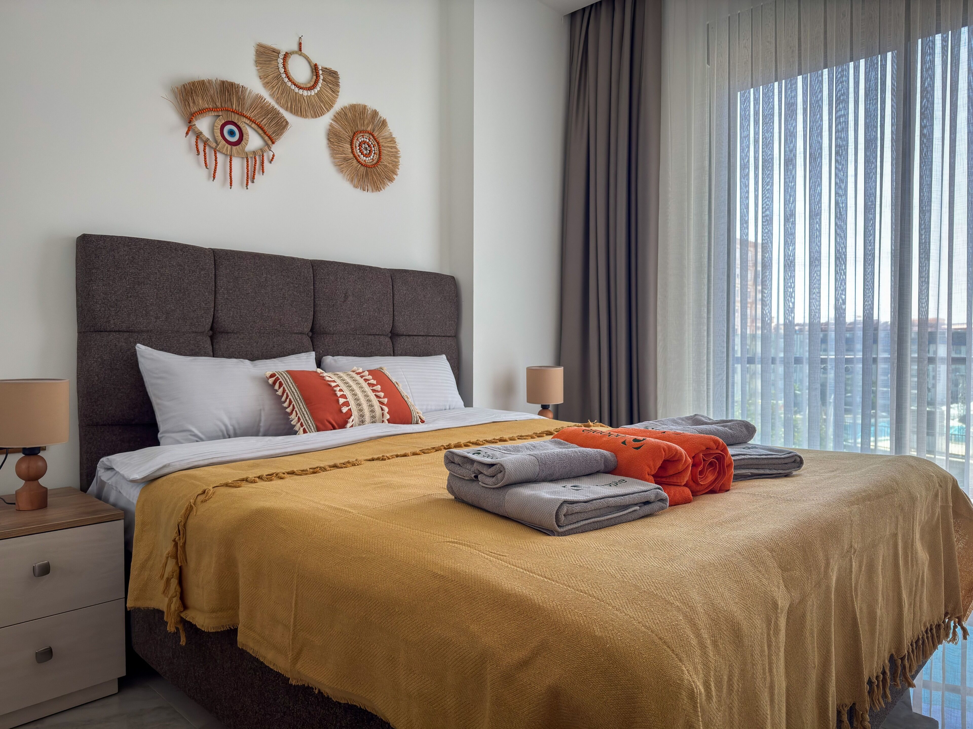 Quarto de hotel com uma cama grande, uma mesa de cabeceira, uma luminária e vista da cidade através de cortinas translúcidas.