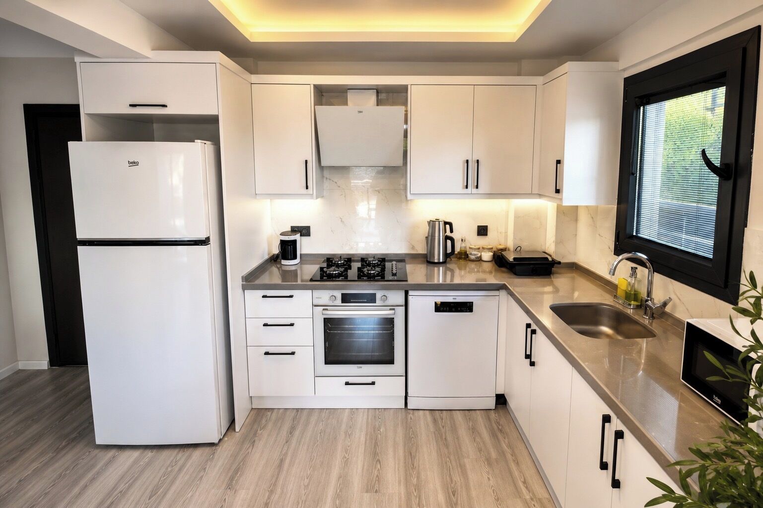 Una cocina moderna con armarios blancos, horno empotrado, refrigerador y una ventana con persianas.