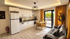 A modern kitchen with white cabinets, a dining table with chairs, and a sofa in a living area with a view to the outside. - Olivara Süite (Muğla)