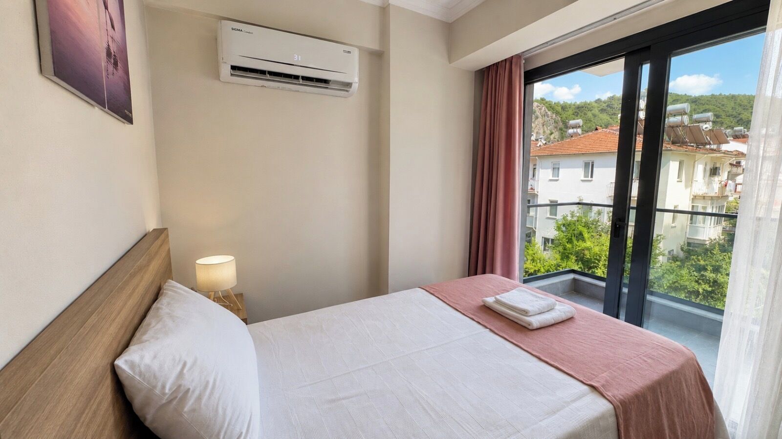 A hotel room with a bed, a nightstand, an air conditioning unit, a window with curtains, and a view of buildings and greenery.