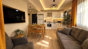 A modern living room with a sofa, a dining table with chairs, a kitchen area with cabinets and appliances, and a large window with curtains. - Olivara Süite (Muğla)