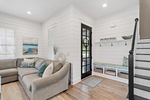 A living room with a beige sofa, a painting of a beach, a black door, and a staircase. - Willow & Waves (Panama City Beach)