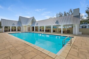 Ein Schwimmbecken mit gefliestem Deck, überdachten Sitzbereichen und blauem Himmel.