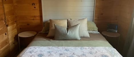 A wooden cabin bedroom with a bed, pillows, a bedside table, and a nightstand.