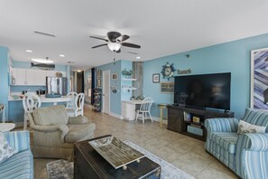 A living room with a television, a ceiling fan, a kitchen in the background, and a dining area. - Seafarer 108- The Peachy Pelican (Destin)