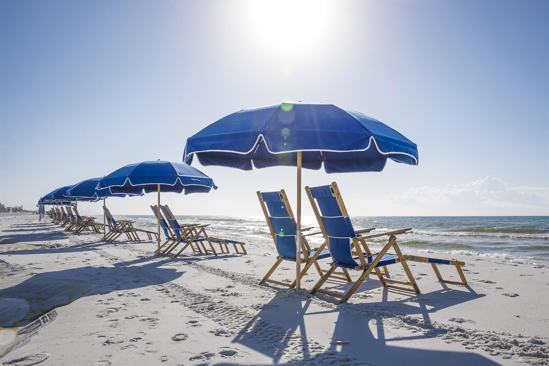 Ein Strand mit blauen Liegestühlen und Sonnenschirmen, Holzliegen und freiem Blick auf das Meer.