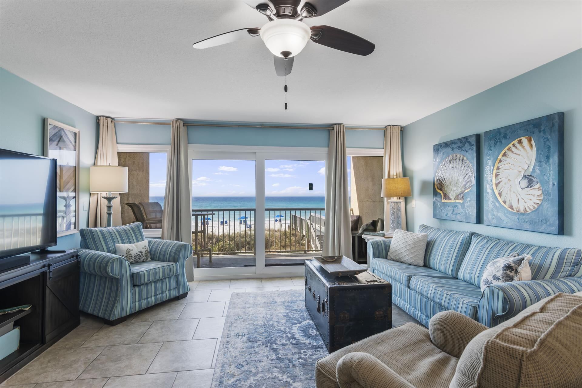 A living room with a television, striped sofas, a coffee table, and a view of the beach.