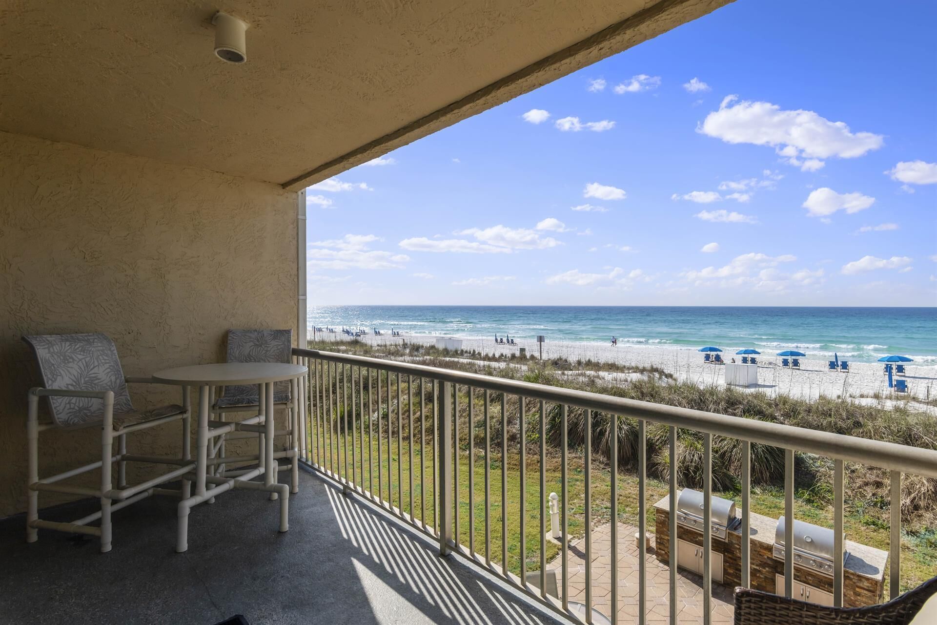 A balcony with a view of the beach, outdoor furniture, and a barbecue grill.