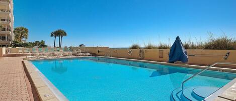 A clear blue pool with a tiled deck, a blue umbrella, and a palm tree in the background.