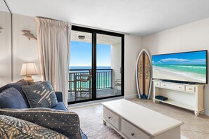 A living room with a television, a surfboard, and a view of the ocean through a sliding glass door. - Sundestin 1405 (Destin)