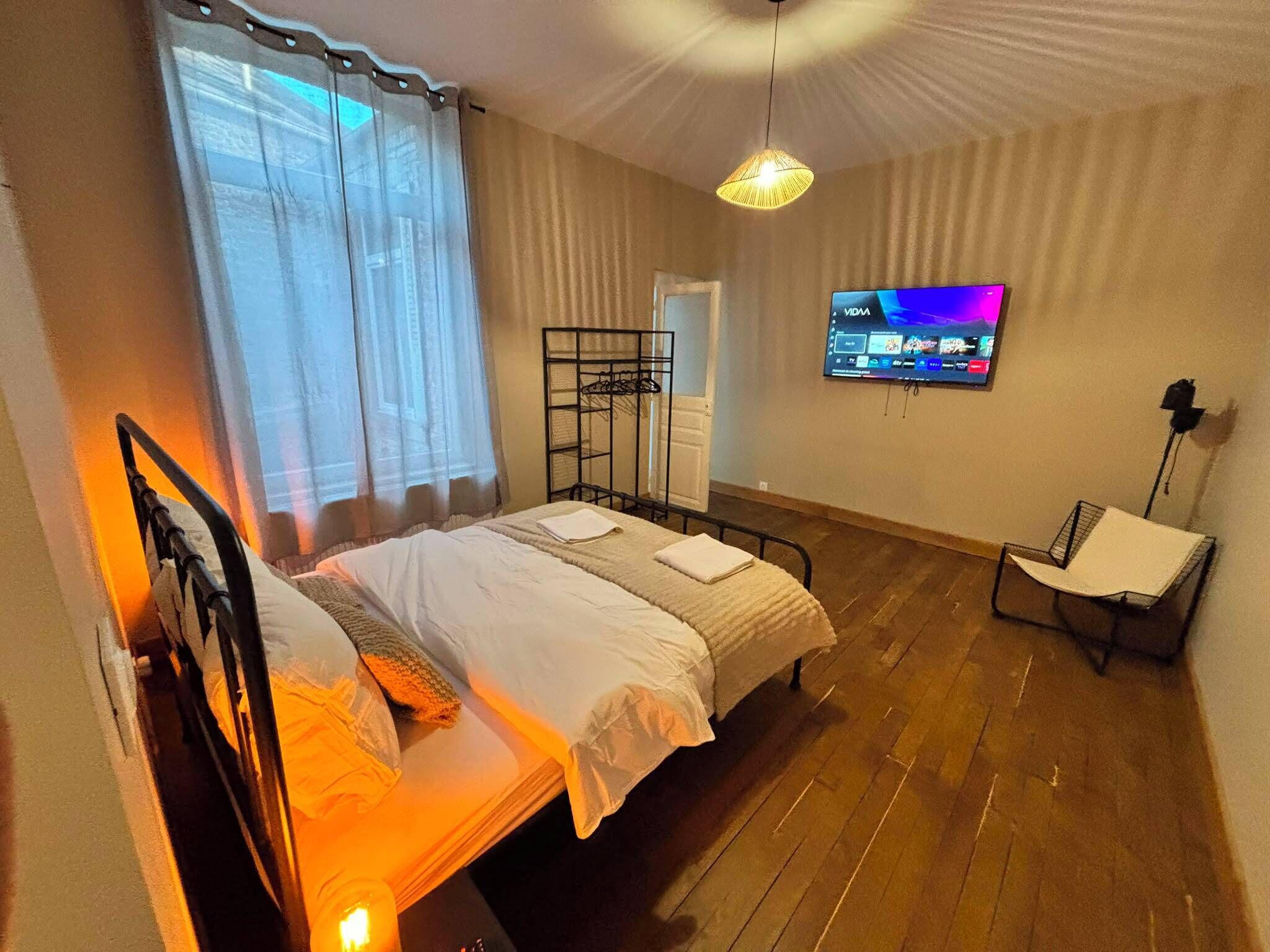 A bedroom with a bed, a TV mounted on the wall, a chair, and a window with curtains.