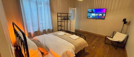 A bedroom with a bed, a TV mounted on the wall, a chair, and a window with curtains.
