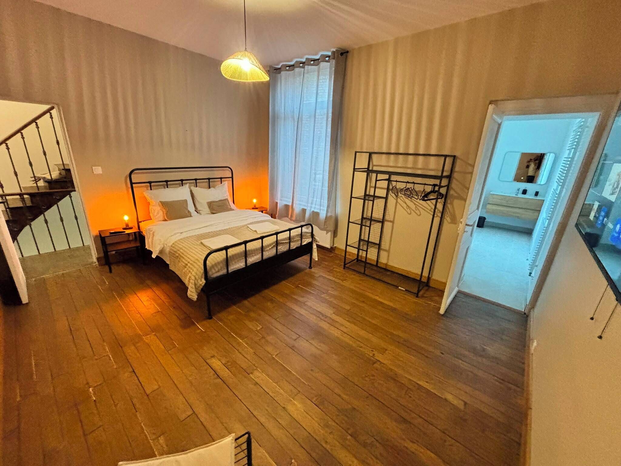 A bedroom with a bed, a nightstand, a hanging light fixture, a window with curtains, a coat rack, and a staircase leading to an upper level.