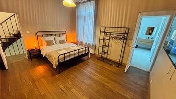 A bedroom with a bed, a nightstand, a hanging light fixture, a window with curtains, a coat rack, and a staircase leading to an upper level.