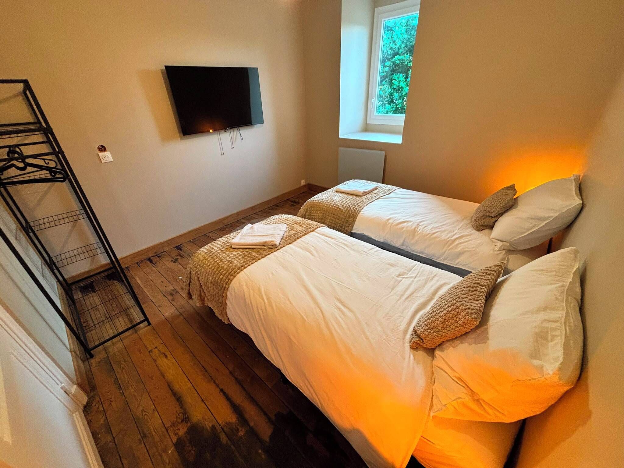 A room with two beds, a TV mounted on the wall, and a window with a view of trees.