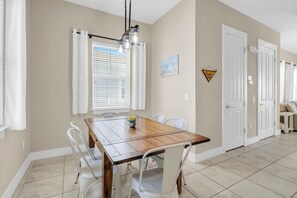 A dining area with a wooden table and white chairs, a window with blinds, and a door with a sign. - Sunny Beach Days (Panama City Beach)