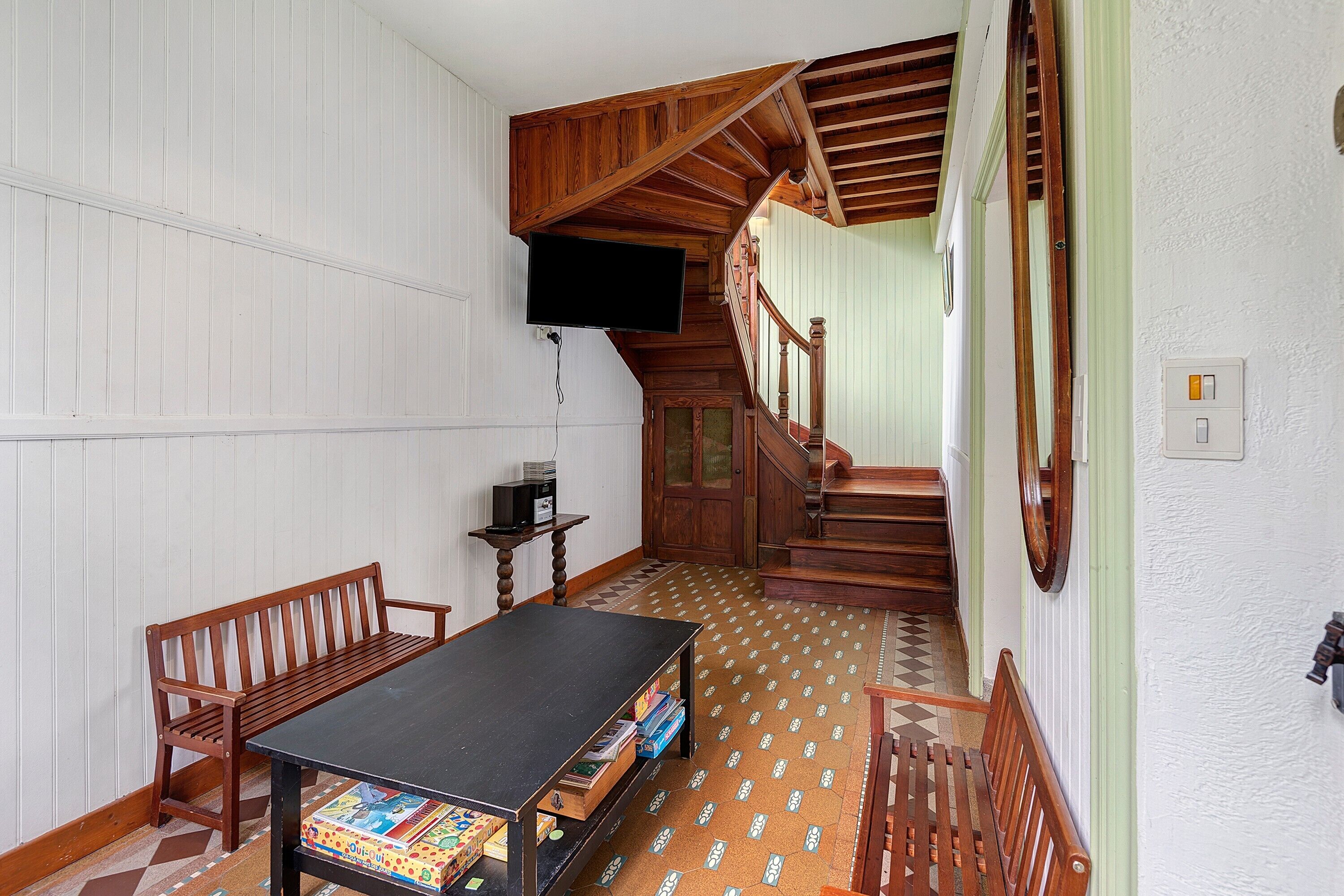 Une pièce avec un escalier en bois, une table noire sur laquelle sont posés des jeux de société et un téléviseur fixé au mur.