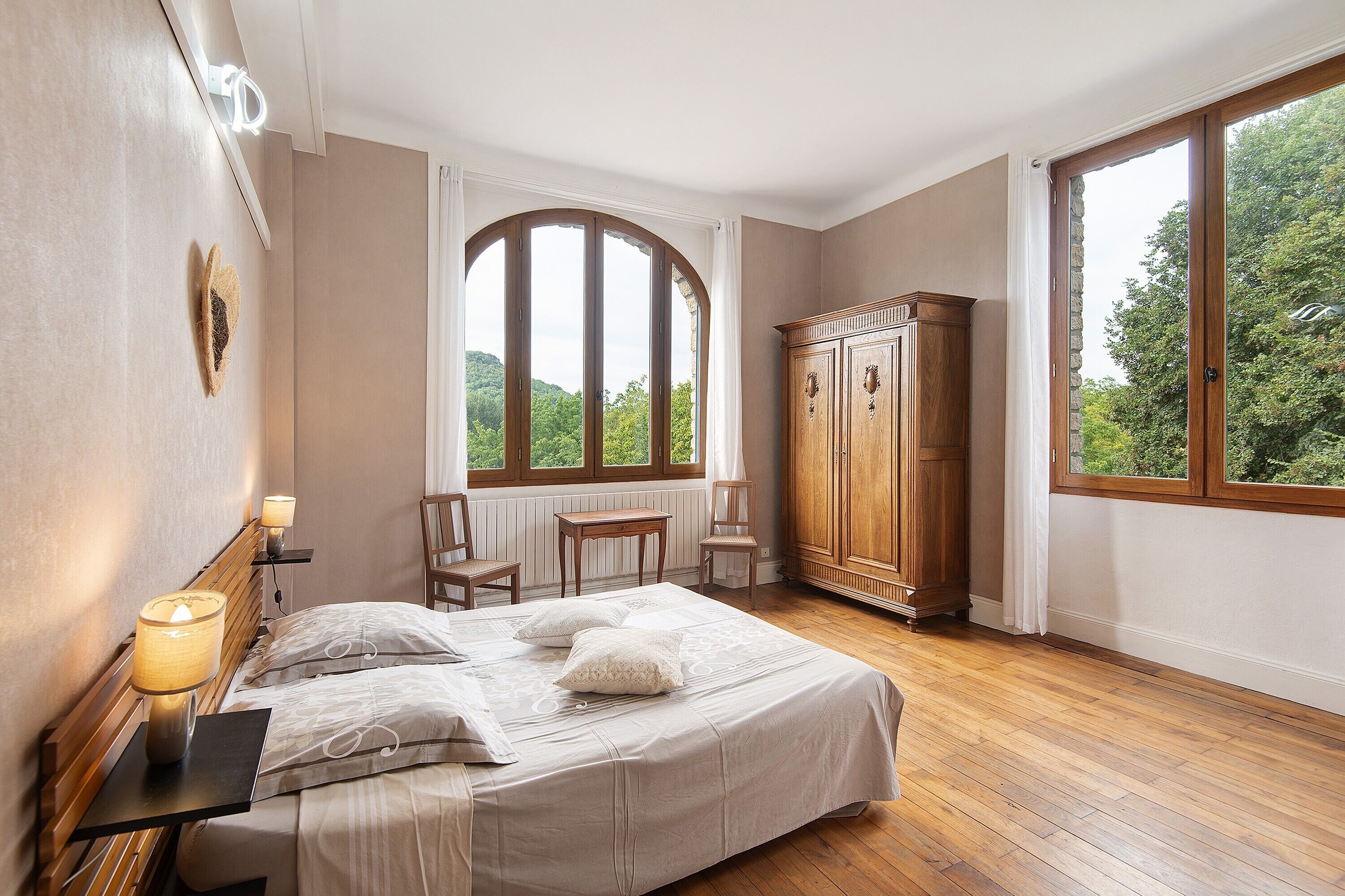 A bedroom with a bed, two chairs, a wooden wardrobe, a window with a view of greenery, and a radiator.