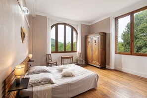 A bedroom with a bed, two chairs, a wooden wardrobe, a window with a view of greenery, and a radiator.