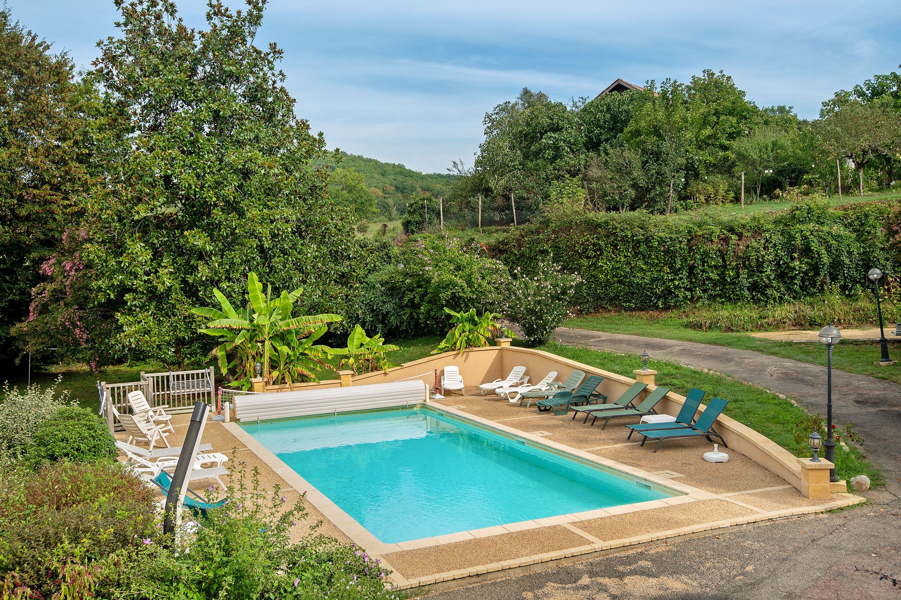 Une piscine rectangulaire bordée de carreaux, entourée d’une terrasse en gravier. On y trouve plusieurs chaises longues et une petite table avec une lampe. La piscine est située dans un espace aménagé avec différentes plantes et arbres.