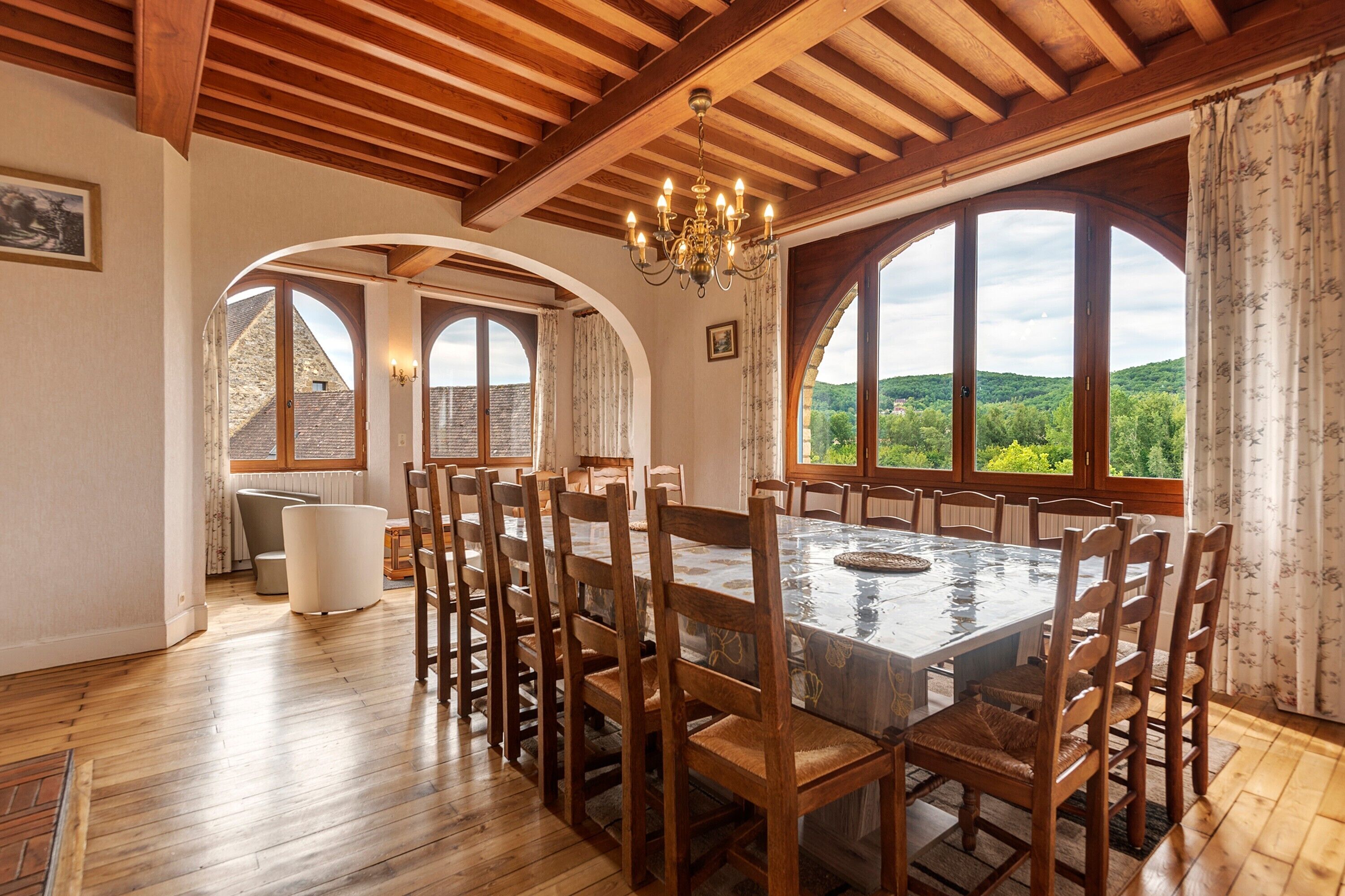 Un espace repas avec une grande table et des chaises en bois, des fenêtres en arc de cercle et un lustre.